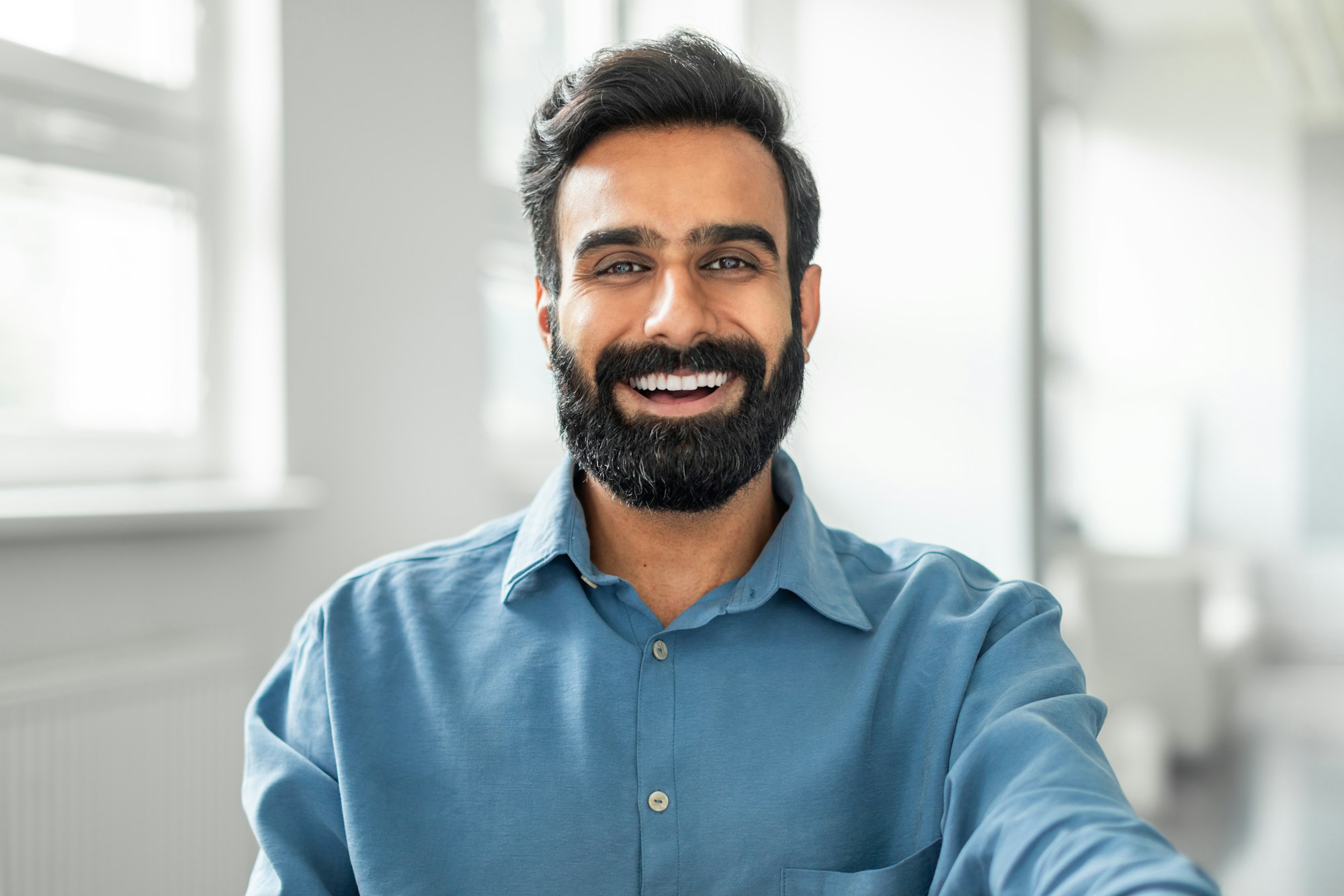 Cheerful smiling indian man wearing shirt sitting at office, closeup photo of happy businessman at
